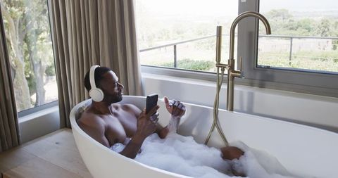 Man relaxing in bubble bath with headphones and smartphone by sunlit balcony window