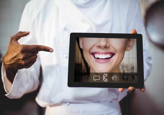 Chef in professional kitchen holding a digital tablet displaying a video call screen with a smiling person. Ideal for use in articles or advertisements about virtual communication in the culinary industry, remote cooking classes, or modern kitchen technology.