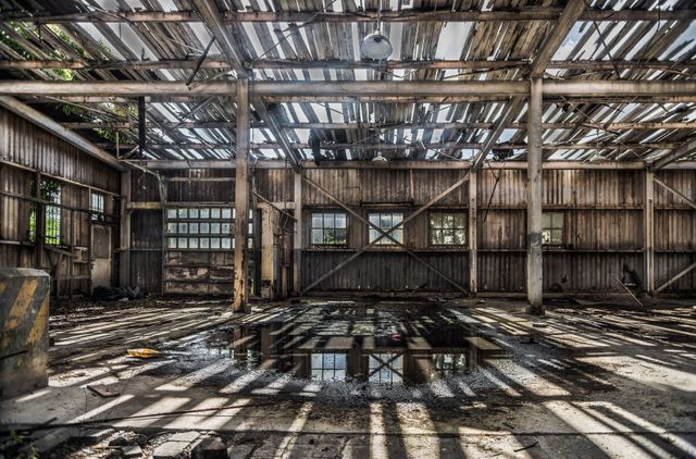 This image depicts an abandoned industrial warehouse with significant decay. The roof is partially collapsed, letting sunlight filter through, creating striking contrasts of light and shadow on the floor. Puddles of water add to the feeling of neglect. Suitable for use in projects about urban exploration, decay, or abandoned places. Ideal for illustrating themes of neglect, decay, and the passage of time in both artistic and commercial contexts.