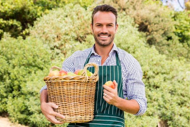 Young man holding a basket of apples while smiling in an orchard. Ideal for use in agricultural promotions, healthy lifestyle campaigns, and gardening blogs. Perfect for illustrating concepts of organic farming, fresh produce, and outdoor activities.