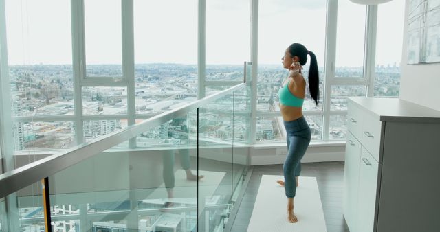 Woman practicing yoga in modern apartment with panoramic windows and city view. Great for content on home workouts, wellness, urban lifestyle, mental health benefits of exercise, and interior design inspiration for living spaces.