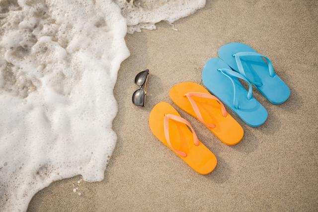 Bright orange and blue flip-flops paired with sunglasses laying on sandy beach near ocean waves. Perfect for themes involving summer vacations, beach holidays, travel, relaxation, and seaside leisure activities.