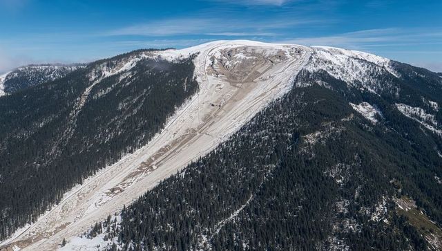 Eroded snow chute carving down alpine ridge, aerial panorama of conifer-covered mountain slopes