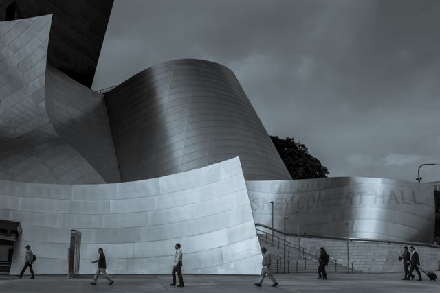 Image shows a modern building with a striking steel facade. Several pedestrians are seen walking past, going about their daily business. The overcast sky adds a moody atmosphere. This photo can be used for urban development projects, architectural showcases, or lifestyle blogs highlighting city life.
