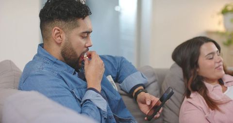 Couple Relaxing on Couch While Man Nibbling Snack and Scrolling Smartphone