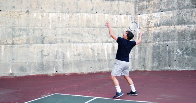 Man holding a tennis racquet serving a ball against a tall concrete wall. He wears athletic clothing including a dark shirt and shorts. Ideal for promoting sports activities, fitness routines, athletic programs, and outdoor practices.