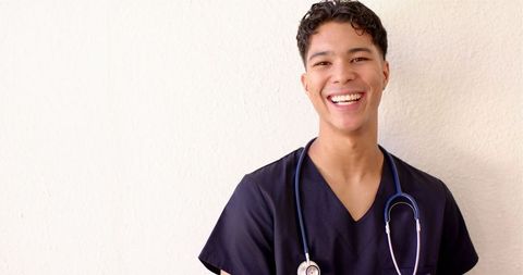 Confident male nurse smiling with stethoscope against light wall