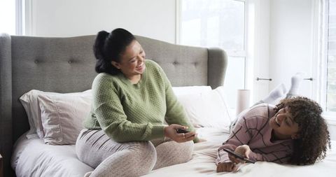 Friends laughing on bed in bright bedroom holding remote and smartphone