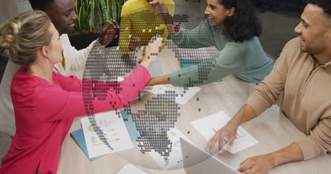 Diverse team joining hands over laptop and charts with dotted globe overlay for global collaboration