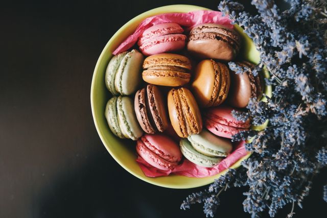 Colorful French macarons presented in a bowl with pink paper lining, next to dried lavender flowers. Each macaron displays a vibrant hue, lending a festive and gourmet appeal. Ideal for illustrating dessert recipes, patisserie websites, food blogs, and culinary magazines.