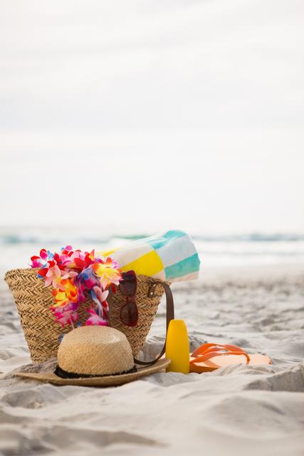 Beach bag filled with summer essentials including a colorful towel, straw hat, sunglasses, and tropical flowers, placed on sandy beach with ocean in background. Ideal for travel blogs, vacation advertisements, summer promotions, and lifestyle magazines.