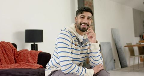 Smiling indian man sitting on sofa in minimalist living room wearing striped sweater