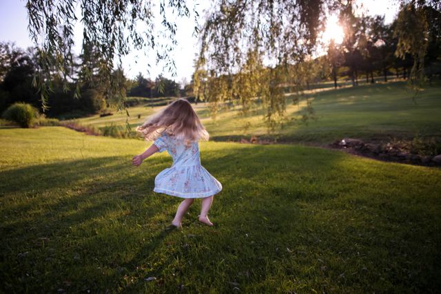 This image portrays a young girl joyfully spinning in a sunlit park. The sun is shining through the trees, casting a warm glow on the grass. Ideal for use in promoting outdoor activities, childhood joy, or summer nature themes.
