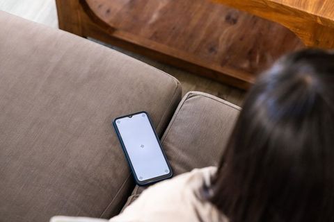 Mature woman sitting on beige sofa turning toward smartphone blank screen for app mockup