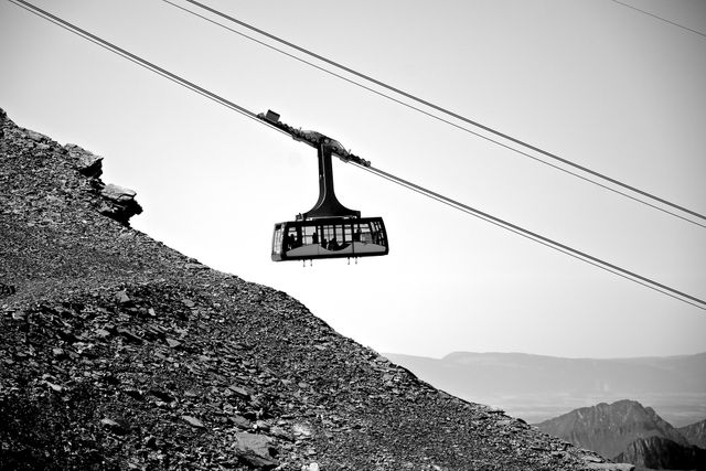 Black and white image of a cable car suspended over a steep rocky slope with mountains in the background. Symbolizes adventure, travel, and high-altitude transport. Perfect for websites and materials related to outdoor activities, mountain excursions, transportation, and adventure tourism.