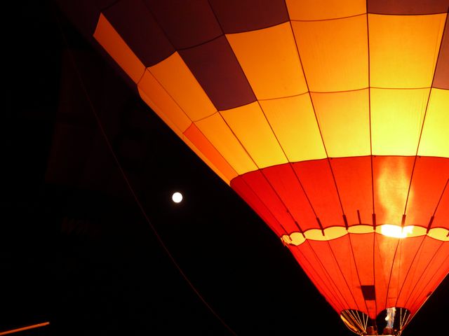 This image depicts a hot air balloon illuminated against a dark night sky with the moon visible. The vibrant colors of the balloon contrast with the darkness, creating a captivating visual. This image can be used for travel brochures, adventure and outdoor activity promotions, or to evoke a sense of wonder and exploration in advertising materials.