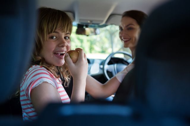 Teenage girl enjoying a healthy snack while sitting in the back seat of a car, with a smiling woman driving. Perfect for illustrating family road trips, healthy eating habits, and joyful travel moments. Can be used in advertisements for family vacations, healthy lifestyle promotions, or automotive services.