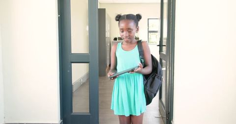 African schoolgirl standing in doorway holding tablet and wearing backpack in mint dress