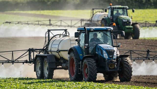 Blue tractor pulling multi-axle sprayer applying crop protection on sunlit arable field