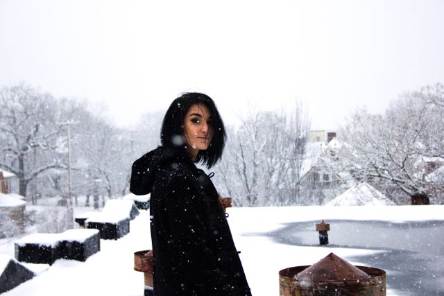 Young woman standing in snowstorm wearing black jacket. Snow covering trees and rooftops in background. Suitable for themes like winter fashion, outdoor activity, season changes, weather conditions.