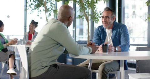 Two coworkers chatting over coffee in modern office lounge with urban cityscape views