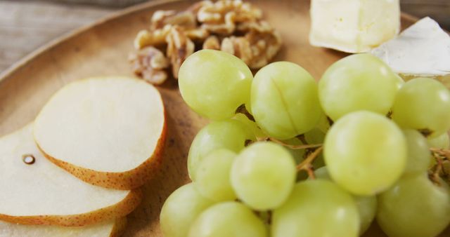 Close-up view of fresh green grapes, slices of pear, walnuts, and pieces of cheese on wooden plate. Perfect for illustrating healthy snacks, fresh food presentations, or nutrition-related content. Can be used in food blogs, recipe websites, and dietary plan materials.