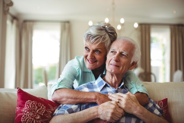 Senior couple embracing while sitting on a sofa in a cozy living room. The couple appears happy and content, showcasing love and togetherness in their retirement years. This image is perfect for use in advertisements, articles, and brochures related to senior living, retirement planning, family life, and home comfort.
