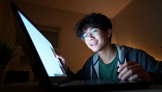 Young man smiling while typing on glowing laptop in dim bedroom workspace at night