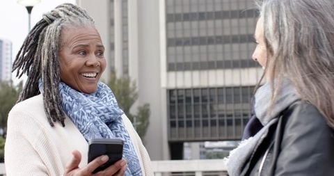 Diverse senior women smiling and chatting on urban bridge, holding smartphone