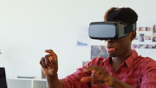 African American male graphic designer using a virtual reality headset, gesturing with hands and smiling in a modern office environment. Ideal for use in articles, blogs, and advertisements related to technology, virtual reality, innovation, workplace creativity, and gaming. Perfect for showcasing the use of VR in professional settings and educational content.