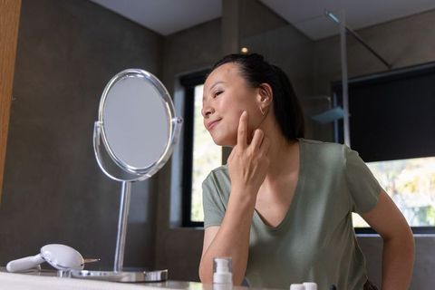 Asian woman practicing skincare routine at bathroom vanity, examining face in mirror