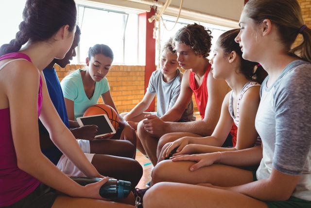 Group of high school students sitting together in a basketball court, using a digital tablet. They appear to be discussing or learning something, possibly related to their sports activity. This image can be used for educational content, sports team promotions, technology in education, and teamwork-related themes.
