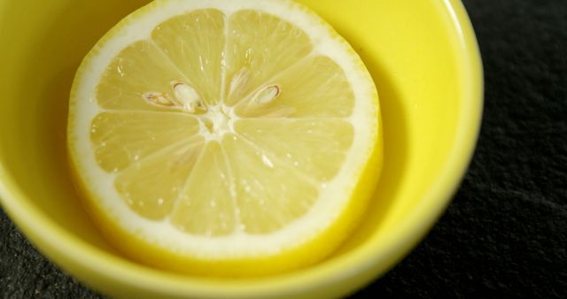 Image shows a close-up of a fresh lemon slice placed in a bright yellow bowl, highlighting the citrus fruit's juicy texture and vibrant color. Suitable for promoting healthy eating, organic food products, food blogs, culinary websites, and nutritional articles.