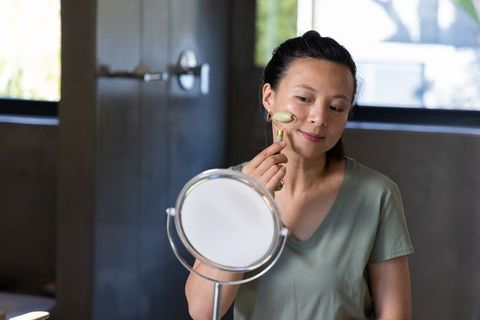 Asian woman using jade roller at bathroom vanity mirror applying facial skincare for glow