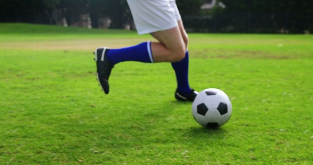 A soccer player in white shorts and blue socks is about to kick a soccer ball on a lush green field, with copy space. Capturing the action mid-motion emphasizes the dynamic nature of the sport and the athlete's skill.