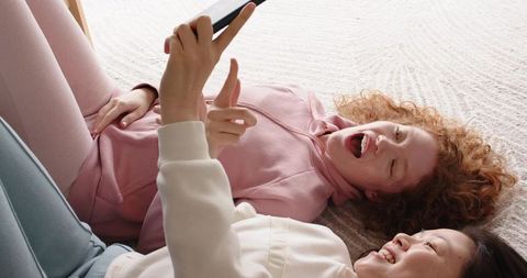 Friends laughing and sharing smartphone while lying on bed in cozy pastel hoodies