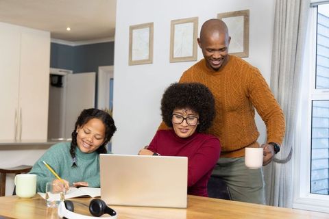 Diverse family working from home at wooden table with laptop and child doing homework