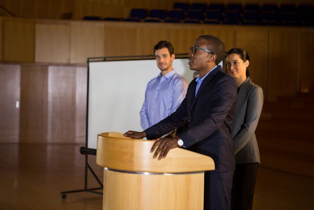 Business executive giving a speech at a conference center with two colleagues standing behind. Ideal for use in articles or advertisements related to business presentations, leadership, corporate events, seminars, and professional development.