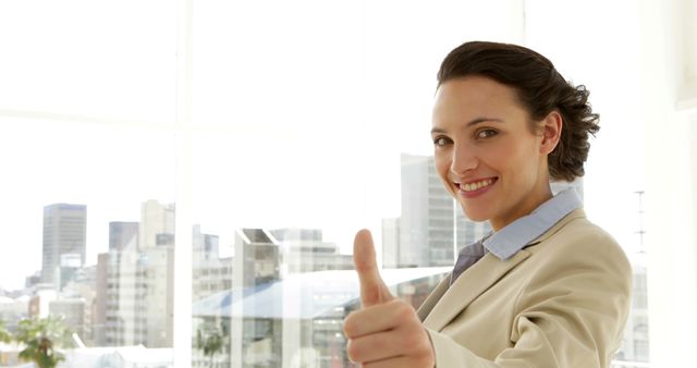 This photo depicts a confident businesswoman standing in a bright office with big windows and a city view, giving a thumbs up. Ideal for use in corporate presentations, business success stories, motivational materials, and professional development content.