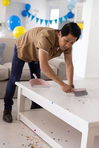 Asian man cleaning confetti from white coffee table after party with balloons and banner
