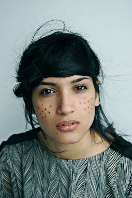 Young woman with face adorned with artistic black dots, standing against neutral background. Ideal for fashion editorials, artistic portraits, beauty brands, creative campaigns, and modern social media content.