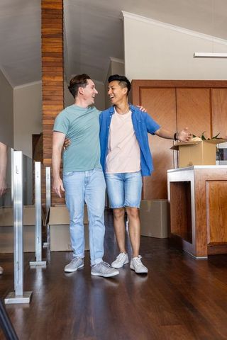 Interracial gay couple moving into new home unpacking boxes walking arm-in-arm and smiling