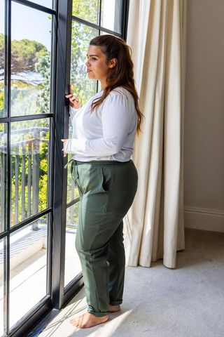Barefoot woman standing at black-framed glass door looking out to sunlit balcony and greenery