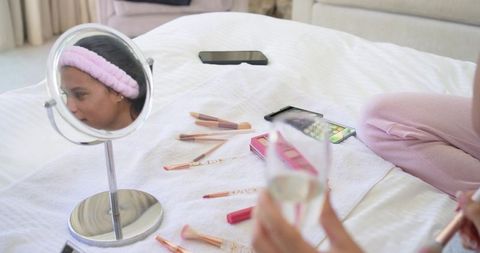 Mother and teen daughter applying makeup on bed with vanity brushes and mirror