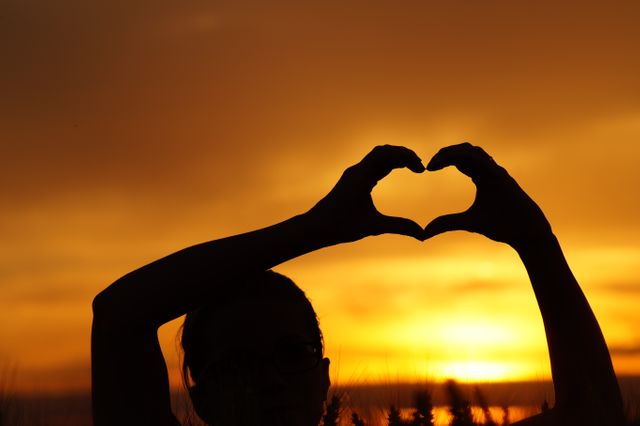 Person forming a heart shape with hands at sunset. Useful in promoting romantic getaways, love and relationships content, natural beauty, and relaxing outdoor activities.