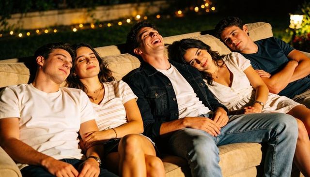 Five friends lounging and laughing on outdoor sofa under string lights