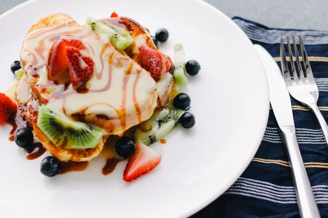 French toast topped with fresh strawberries, kiwi, and blueberries, drizzled with syrup on white plate, paired with a fork and knife next to a cloth napkin. Ideal for food blogs, recipe websites, breakfast and brunch promotions, and restaurant menus.