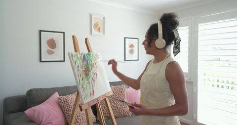 African american woman painting abstract canvas in cozy living room with headphones