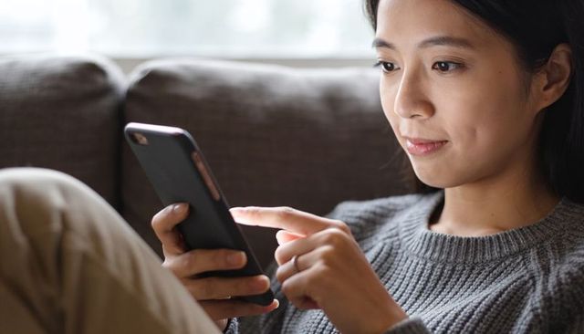 Young woman scrolling smartphone on sofa tapping screen wearing ring relaxed at home