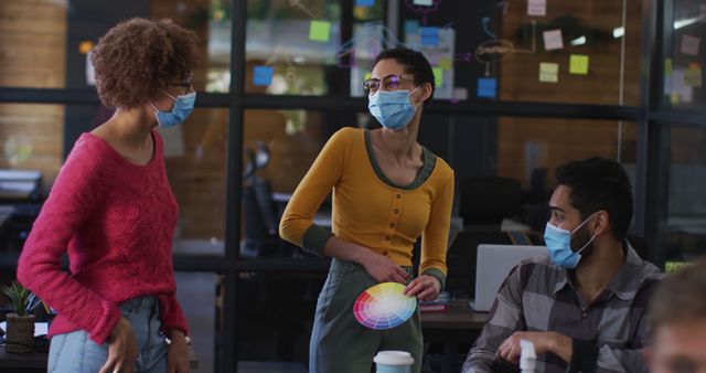 Group of professionals working together in modern office environment during pandemic. Wearing face masks and socially distancing while engaging in discussions, holding color samples, and sharing ideas. Ideal for use in content related to creative industry, workplace safety, COVID-19 precautions, and teamwork.